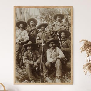 May include: A vintage black and white photograph of a group of seven men wearing hats and holding rifles. The men are standing in a field and appear to be dressed in traditional clothing.
