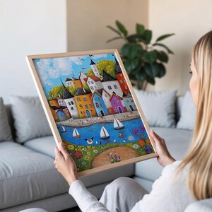 Puede incluir: Una pintura enmarcada de un pueblo costero colorido con casas, barcos y flores. La pintura est&aacute; en un marco de madera claro.
