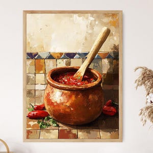Op de afbeelding: Een ingelijst schilderij toont een rustieke terracotta pot gevuld met rode saus en een houten lepel. Rode chilipepers en groene kruiden zijn rond de pot verspreid. De achtergrond heeft een betegelde muur met een neutraal kleurenpalet.