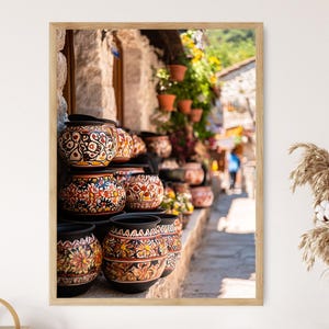May include: A stack of colorful, hand-painted ceramic pots with intricate designs. The pots are arranged in a shop window, with a view of a cobblestone street in the background.