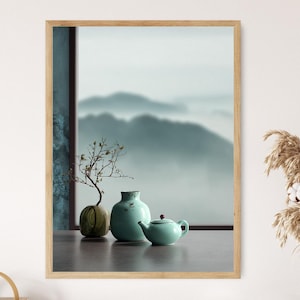 May include: A framed print featuring a serene, misty mountain landscape. The foreground displays a small, light green teapot, a turquoise vase, and a round, olive-green decorative object. A vase of dried flowers is on the right.