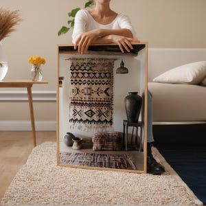 May include: A framed display featuring a woven textile with geometric patterns in black, beige, and red. The display includes a small rug, a dark vase, and a hanging lamp. The scene is set on a beige rug.