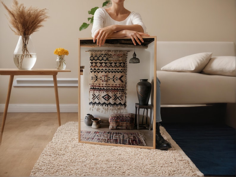 May include: A framed display featuring a woven textile with geometric patterns in black, beige, and red. The display includes a small rug, a dark vase, and a hanging lamp. The scene is set on a beige rug.