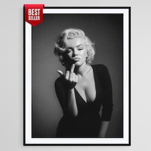 Puede incluir: Impresión enmarcada en blanco y negro que presenta a una mujer con cabello rubio, vestida con un vestido negro con escote. La mujer hace un gesto grosero con el dedo medio. Una etiqueta roja de "Best Seller" está en la esquina superior izquierda.