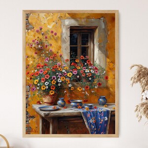 Op de afbeelding: Een schilderij van een geel gebouw met een raam en een bloembak vol met kleurrijke bloemen. De bloemen vallen uit de bak en op een tafel met blauw-witte borden en een blauw tafelkleed.