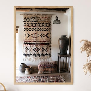 May include: A framed mirror reflects a bohemian-style interior. A patterned wall hanging, a large dark vase on a small stool, and a patterned rug are visible. The color palette includes beige, black, and brown.