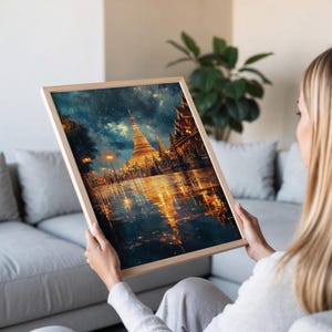 May include: A framed art print depicting a golden pagoda and other buildings reflecting in a body of water. The artwork has a painterly style with a dark, moody sky and warm, glowing lights. The frame is made of light-colored wood.