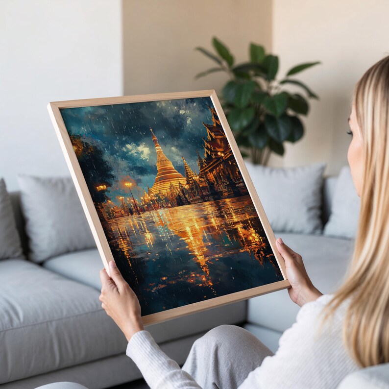May include: A framed art print depicting a golden pagoda and other buildings reflecting in a body of water. The artwork has a painterly style with a dark, moody sky and warm, glowing lights. The frame is made of light-colored wood.