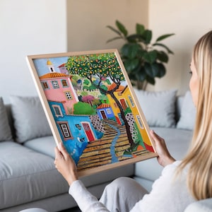 May include: A framed colorful painting depicting a European village scene. The artwork features buildings in shades of blue, pink, and yellow, with a tree and a winding path. The frame is made of light-colored wood.