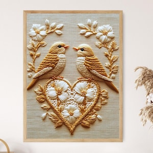 May include: Embroidered artwork featuring two birds facing each other, perched on a heart-shaped design. The artwork is framed in a light wood frame. The birds and heart are in shades of gold and white, with floral accents.