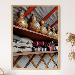 May include: A framed image showcases a wooden shelf filled with decorative gourds, each adorned with intricate carvings. Below, stacked textiles and metal cups are visible. The scene is set within a rustic interior, with a wooden frame.