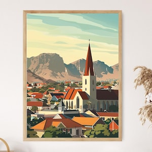 Op de afbeelding: Een ingelijste kunstdruk die een gestileerd stadsbeeld toont met een kerk en rood bedekte gebouwen. Het kunstwerk toont een hoge kerk met een rode torenspits, tegen een achtergrond van bergen en een blauwe lucht. De print zit in een houten lijst.