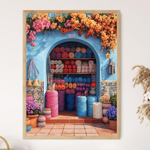 Op de afbeelding: Een ingelijste print van een wolwinkel met een blauwe boogvormige ingang, gevuld met kleurrijke garenbollen. Roze en oranje bloemen sieren de bovenkant. De buitenkant van de winkel heeft potplanten en decoratieve tegels.
