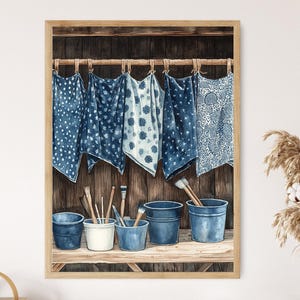 Op de afbeelding: Waterverfschilderij van een rustieke werkplaats. Vijf blauw-wit patronen doeken hangen aan een houten roede. Daaronder staan ​​verschillende emmers en penselen op een houten plank, wat een charmante, artistieke weergave creëert. Het kleurenpalet wordt gedomineerd door blauw- en bruintinten.