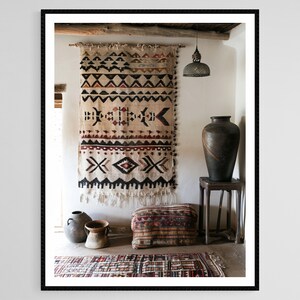 May include: A framed interior shot features a beige wall hanging with geometric patterns in black, red, and white. A large, dark vase sits on a wooden stool. Other decorative items include a patterned ottoman and a small rug.