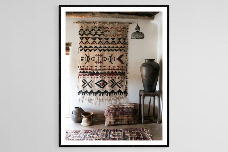 May include: A framed interior shot features a beige wall hanging with geometric patterns in black, red, and white. A large, dark vase sits on a wooden stool. Other decorative items include a patterned ottoman and a small rug.