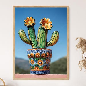 Könnte beinhalten: Gerahmter Druck eines farbenfrohen Keramikkaktus in einem dekorativen Topf. Der Kaktus ist grün und gelb mit orangefarbenen Blüten. Der Topf ist blau mit orangefarbenen Blumenmustern. Der Hintergrund ist ein klarer blauer Himmel.