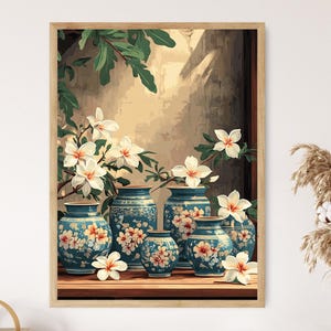 May include: A framed print featuring a still life of blue and white ceramic vases with floral designs. White flowers with yellow centers and green leaves are arranged around the vases. The background has a window and foliage.