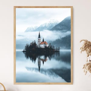 May include: A framed print of a serene landscape featuring a church on a small island in a lake. The church has a red roof and a tall steeple. The water reflects the church and the surrounding mountains, creating a symmetrical image. The sky is overcast.