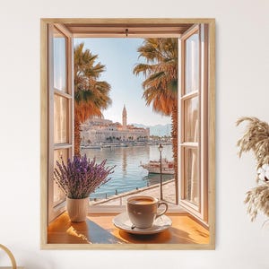 Op de afbeelding: Een ingelijste prent van een schilderachtig uitzicht door een open raam. Het raam onthult een kustplaats met palmbomen, een kerktoren en boten op het water. Een kopje koffie op een schoteltje staat op de vensterbank, naast een vaas met lavendel.