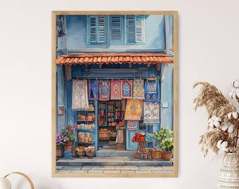 May include: A framed painting of a storefront with blue walls and a terracotta tiled awning. The shop displays various colorful patterned textiles hanging in the entrance, with potted plants and baskets of goods arranged on the stone sidewalk.