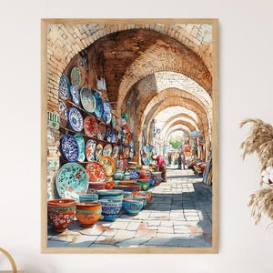 May include: Watercolor painting of a market scene with arched brick walkways. The walls are lined with colorful ceramic plates and bowls. The artwork is framed in a light wood frame.