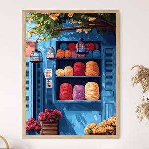 Op de afbeelding: Een ingelijste print van een levendige garenwinkel. Het kunstwerk toont een blauwe gevel met planken vol kleurrijke bollen garen. Decoratieve lantaarns hangen van bovenaf en bloemen sieren de bloembakken, wat een vrolijke sfeer creëert.