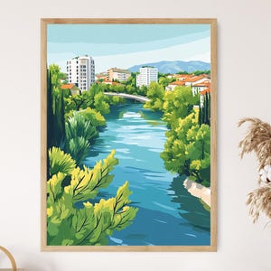 Könnte beinhalten: Ein gerahmter Kunstdruck mit einer Stadtlandschaft mit einem Fluss. Das Kunstwerk zeigt Gebäude, Bäume und eine Brücke, mit einer Farbpalette aus Blau-, Grün- und Orangetönen. Der Druck ist in einem Holzrahmen.