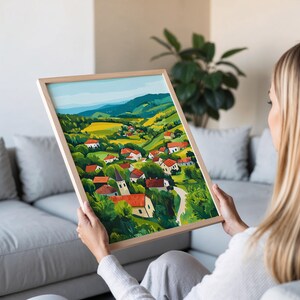 May include: A framed painting of a village nestled in a valley with rolling green hills and a blue sky. The village is made up of small houses with red roofs and a church steeple.