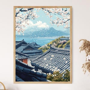 May include: A framed art print depicting a Japanese landscape. The artwork features traditional buildings with dark blue tiled roofs, cherry blossoms, and a mountain in the background. The sky is a light blue, and the overall scene evokes a sense of tranquility.