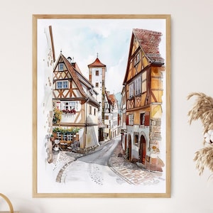 May include: Watercolour painting of a European town street scene. The artwork features detailed buildings with red-tiled roofs, timber-framed walls, and cobblestone streets. The painting is framed in a light-coloured wooden frame.