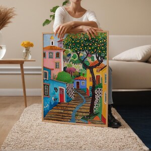 May include: A colorful framed painting depicts a European village scene. The artwork features buildings in shades of pink, blue, and yellow, with a tree bearing fruit. A stone staircase leads up a hill. The painting is held by a person.