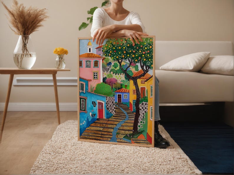 May include: A colorful framed painting depicts a European village scene. The artwork features buildings in shades of pink, blue, and yellow, with a tree bearing fruit. A stone staircase leads up a hill. The painting is held by a person.