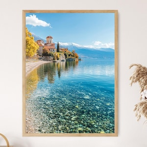 May include: A framed print of a scenic landscape featuring a church with a red-tiled roof on a rocky shoreline. The clear blue water reflects the sky and surrounding mountains. The image is framed in a light-colored wooden frame.