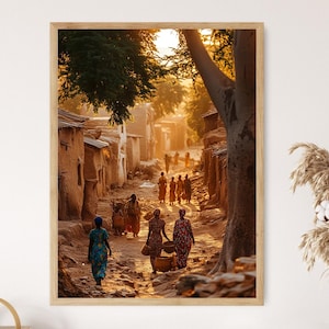 May include: A framed print of a sunlit village scene. The image shows a narrow dirt road lined with mud-brick buildings. People in colorful clothing walk along the road, some carrying baskets. The warm light creates a golden glow.