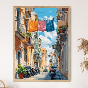 Könnte beinhalten: Ein gerahmter Kunstdruck zeigt eine sonnige italienische Straßenszene. Bunte Gebäude säumen die schmale Straße, mit Wäsche, die darüber hängt. Roller parken am Bürgersteig, und Topfpflanzen fügen Grün hinzu. Der Himmel ist ein helles Blau mit flauschigen weißen Wolken.