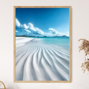 May include: Framed print of a tropical beach scene. The image features a pristine white sand beach with rippled patterns, turquoise water, and a bright blue sky with fluffy white clouds. The artwork is encased in a light brown wooden frame.