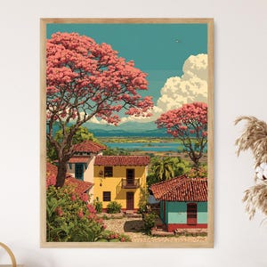 May include: A framed art print depicting a vibrant landscape with colorful buildings and lush pink flowering trees. The artwork features a bright blue sky with fluffy white clouds, a body of water, and distant mountains. The buildings have red tile roofs.