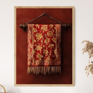 Op de afbeelding: Een rood wandkleed met gouden bloemenpatronen hangt aan een houten stok tegen een rode achtergrond. Het wandkleed heeft een franje rand.