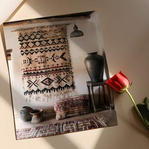 May include: A photograph of a room interior featuring a beige and black patterned wall hanging, a dark vase on a small wooden table, and a red tulip. Other items include a patterned ottoman and small pottery.