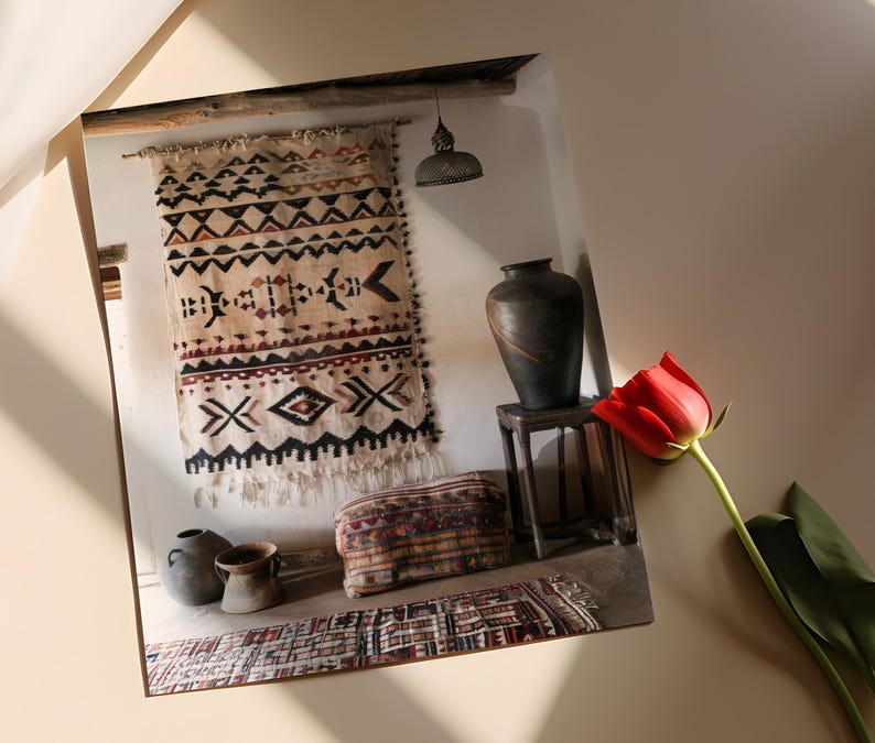 May include: A photograph of a room interior featuring a beige and black patterned wall hanging, a dark vase on a small wooden table, and a red tulip. Other items include a patterned ottoman and small pottery.