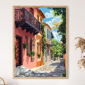 May include: A framed print showing a picturesque street scene. The artwork features buildings with pink and orange walls, balconies, and a cobbled street. The scene is enlivened by greenery and a bright blue sky with clouds.
