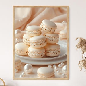 May include: A framed print featuring a stack of cream-colored macarons on a white plate, surrounded by pearls. The macarons have a textured appearance and are arranged in a visually appealing composition. The background includes soft, draped fabric.