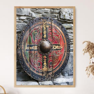 May include: A framed print of a round, wooden Viking shield. The shield features a central boss and is decorated with red, green, and gold Celtic knotwork designs. The shield is mounted against a stone wall.