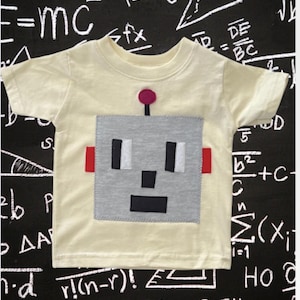 May include: A light yellow short-sleeved t-shirt featuring a gray robot face applique. The robot has black and white details, red accents, and a pink antenna. The background is a chalkboard with mathematical equations.