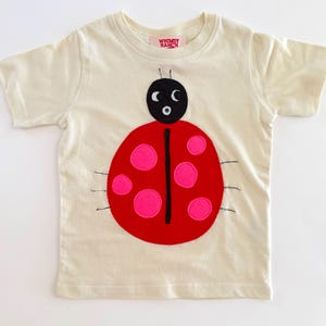 May include: A cream-colored short-sleeve t-shirt featuring a large ladybug appliqué on the front. The ladybug has a black head with white eyes, a red body with bright pink circular spots, and thin black embroidered legs. The label reads Whimsy Ranch.