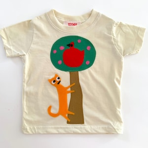 May include: A cream-colored short-sleeve t-shirt featuring a central applique design of a brown tree trunk with a green canopy containing pink flowers and a red bird. An orange cat is positioned climbing the tree trunk. The label reads Whimsy.