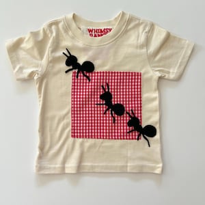 May include: A cream-colored short-sleeve t-shirt featuring a square red and white gingham patch stitched onto the front. Three black ant silhouettes are appliqued across the shirt, with one positioned on the patch and two others nearby.