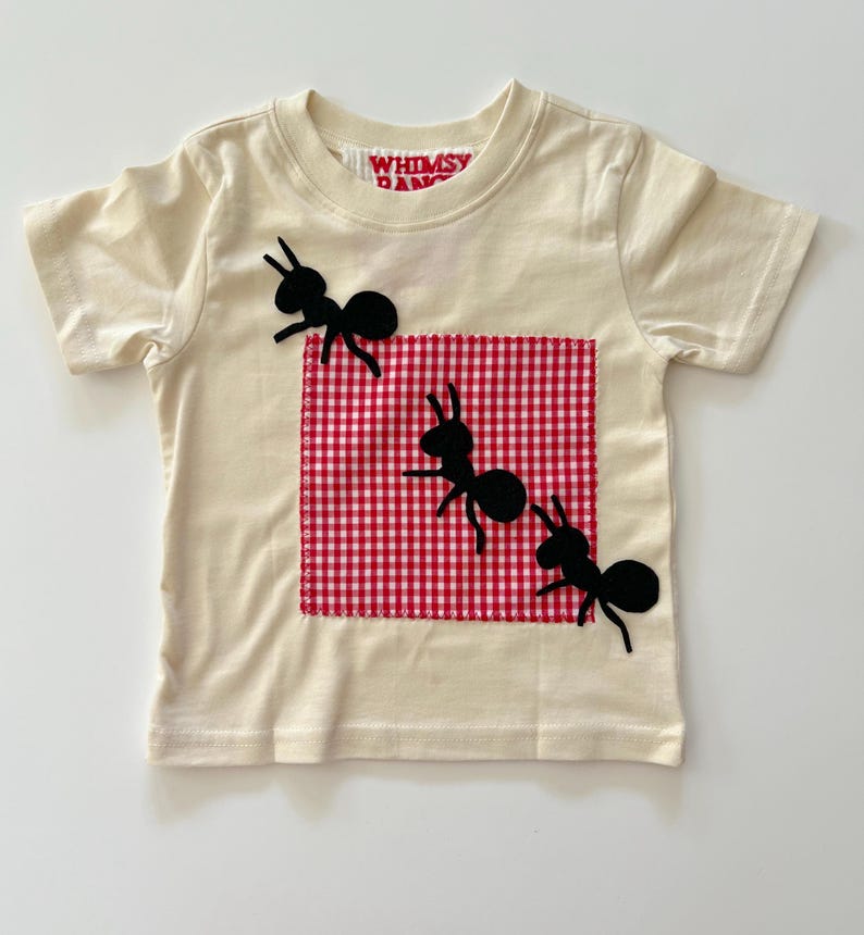 May include: A cream-colored short-sleeve t-shirt featuring a square red and white gingham patch stitched onto the front. Three black ant silhouettes are appliqued across the shirt, with one positioned on the patch and two others nearby.