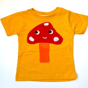 May include: A short-sleeved mustard yellow t-shirt featuring a large, centered applique of a red mushroom with a bright orange stem. The mushroom cap has a smiling face with two black eyes and three white spots, creating a playful, graphic design.